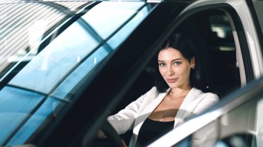 Kendine güvenen bir kadın bir galeride arabada oturuyor, aracın içini ve şık tasarımını vurguluyor, araba satın alma yolculuğunu somutlaştırıyor.