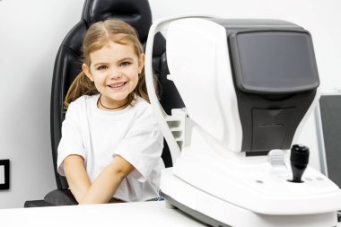 Gülümseyen kız göz muayenesi ekipmanlarının yanında oturuyor.