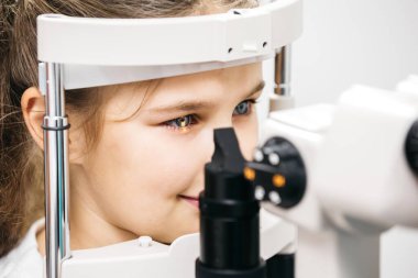 Genç kız ileri göz teçhizatıyla göz muayenesine odaklanarak, klinik bir ortamda düzenli görme kontrolleri ve pediatrik göz sağlığının önemini vurguluyor