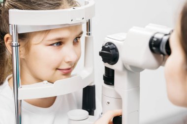 Genç kız, göz muayenesinde uzmanlaşmış göz teçhizatı kullanılarak pediatrik göz bakımının önemini ve erken görüş değerlendirmelerine duyulan ihtiyacı vurgulamaktadır.