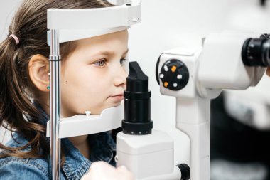 Genç bir kız ileri düzey göz teçhizatıyla göz muayenesine katılıyor. Klinik bir ortamda pediatrik göz bakımının önemini vurguluyor.