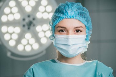 Ameliyat maskesini takmış genç bir kadın cerrah ameliyathanede duruyor. Parlak cerrahi ışıklarla aydınlatılıyor. Profesyonelliğini ve ameliyata hazır olduğunu gösteriyor.