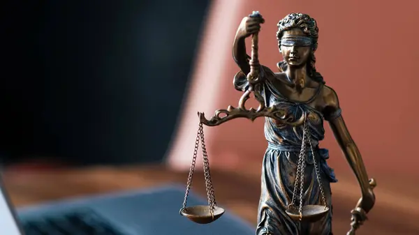 Adalet Anıtı 'nın terazili bronz heykeli, bir dizüstü bilgisayarın yanındaki ahşap masaya yerleştirilmiş, hukukun, adaletin ve hukuk mesleğinin özünü somutlaştırıyor.