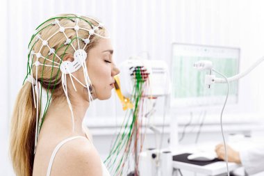 Kafasında elektrotlarla, klinik ortamda EEG incelemesine katılan kadın, tıbbi teknoloji ve hasta bakımını ön plana çıkarıyor.