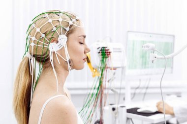 Kafasında elektrotlarla sağlık ortamında EEG testi yaptıran kadın, ekipmanları izlerken beyin aktivitelerini yakalıyor, sağlık hizmetlerindeki yenilikleri vurguluyor.