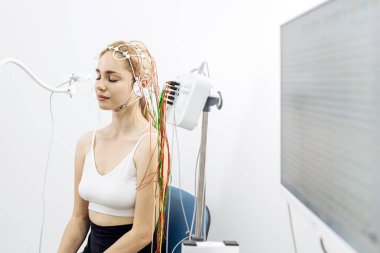 Uzun saçlı bir kadın hasta klinik bir ortamda oturuyor, EEG testine tabi tutuluyor. Kafasına kablolar bağlı ve beyin aktivitesini gösteren bir monitör var.