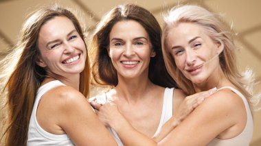 Farklı etnik kökenlere sahip üç kadın sağlıklı bir spa ortamında neşeli bir şekilde kucaklaşıyorlar, parıldayan derilerini ve sağlıklı saçlarını vurguluyorlar, rahatlatıcı dokular ve dingin bir ortam ile çevrili.