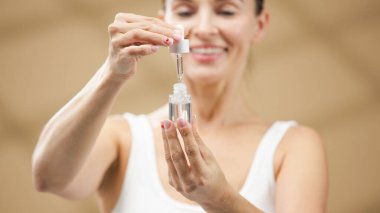 Kadın elinde damlalık şişesi, dikkatle sunulan cilt serumu, yumuşak dokular ve sıcak ışıklandırma ile güzellik rejiminin altını çizmek, refah ve kişisel bakım uygulamaları için rahatlatıcı bir atmosfer yaratmak.