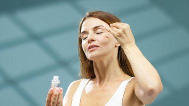 Kadın model yüz serumunu bir damlatıcıyla nazikçe uyguluyor, sakin bir sağlık ortamında rahatlatıcı bir cilt bakımı ayinini vurguluyor, hidrasyon ve parlayan cilt yenilenmesine odaklanıyor.