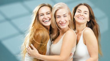 Değişik saç renkleri ve stilleri olan üç kadın sağlıklı bir spa ortamında neşeli bir şekilde kucaklaşıyorlar, rahat, yumuşak, doğal ışıkla dolu bir yerde bir topluluk ve mutluluk hissi sergiliyorlar.