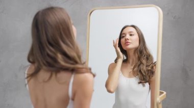 Genç bir kadın aynaya bakarken yüzüne dokunuyor.