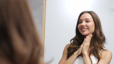 Gülümseyen kadın aynaya bakarken boynuna dokunuyor.