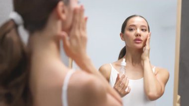 Genç bir kadın aynaya bakarken serum uyguluyor.