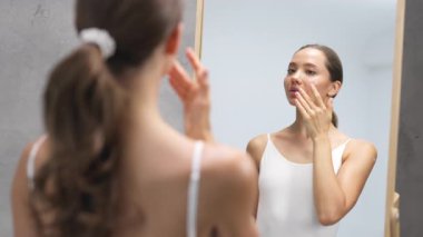 Genç bir kadın aynaya bakarken yüzüne dokunuyor.