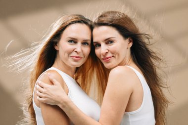 Işıl ışıl cildi ve uzun saçları olan iki kadın, yumuşak bir ortamda birbirlerini kucaklıyor, doğal güzelliklerini, cilt bakımlarını ve sağlıklarını uyumlu bir ortamda ön plana çıkarıyorlar.
