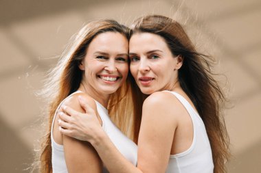 Uzun saçlı iki kadın neşeli bir kucaklaşmayı paylaşıyor. Parlak ve havadar bir ortamda sağlıklı bir cilt ve sağlık yayıyorlar. Cilt bakımı, arkadaşlık ve destek bağının önemini vurguluyorlar.