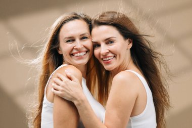 Beyaz kolsuz tişörtlü, uzun saçlı iki neşeli kadın birbirlerini kucaklıyor, neşe ve sağlıklı cilt yayıyor, sıhhat ve cilt için ideal sıcak bir atmosfer yaratıyor ve fotokopi alanı ile cilt teması oluşturuyor.