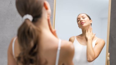 Uzun saçlı bir kadın, sağIıklı cildine bir aynanın önünde hayran, boynuna nazikçe cilt ürünleri uyguluyor, refahı ve kendine bakımı vurgulayan sakin bir atmosfer yaratıyor.