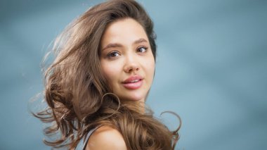 Uzun saçlı genç bir kadın, parlak cildini ve doğal güzelliğini sergiliyor. Yumuşak mavi bir arka planda duruyor. Sağlıklı cilt bakımının ve dingin bir güzellik ortamının önemini vurguluyor.