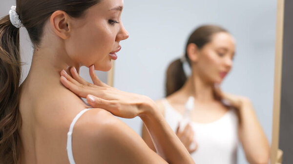 Молодая женщина нежно наносит продукт по уходу за кожей на шею, глядя в зеркало, подчеркивая свою приверженность здоровой рутины кожи и важность ухода за собой в спокойной ванной комнате