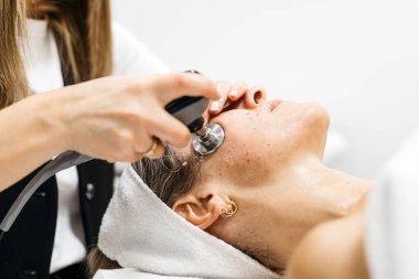Dişi müşteri, cilt dokusunu geliştirmek ve sakinleştirici spa ortamındaki güzelliği artırmak için bir cihaz kullanan bir teknisyenle yüz gençleştirme için RF kaldırma prosedüründen geçiyor.