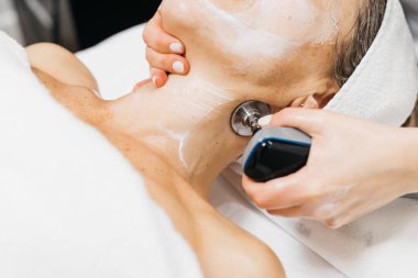 Yüz gençleştirme için RF kaldırma prosedürüne tabi tutulan bayan müşteri, boynunda bir cihaz bulunan bir teknisyen, cilt bakımı ve gevşeme ortamını ön plana çıkarıyor.