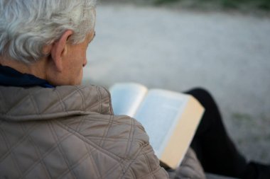 Yaşlı adam, gri saçlı, parkta bir bankta oturmuş kitap okuyor.