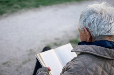Yaşlı adam, gri saçlı, parkta bir bankta oturmuş kitap okuyor.