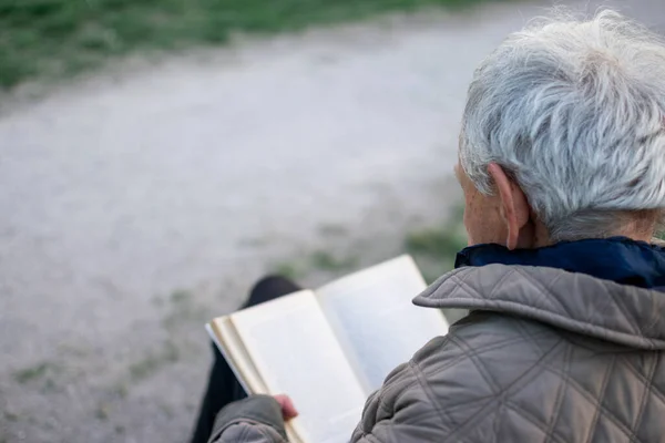 stock image Old man with gray hair reading a book sited on a bench outside in park