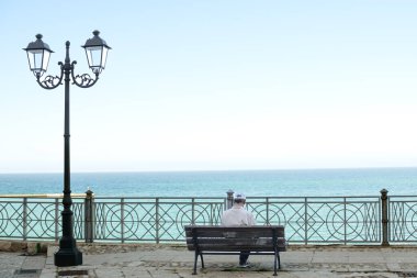 Beyaz bardaklı ve ceketli son sınıf öğrencisi bankta oturmuş deniz altyapısı olan kitapları okuyor.
