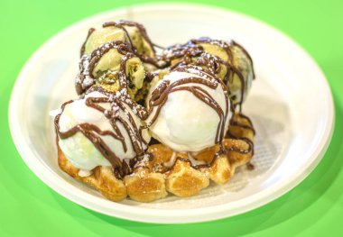 Açık yeşil arka planda beyaz plastik bir tabakta waffle 'ın üzerine serpiştirilmiş İtalyan fıstıklı dondurma.
