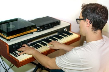 Gözlüklü, beyaz tişörtlü, piyano besteleyen ve fotokopi aletiyle piyano çalan genç bir adam.