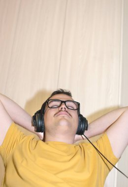 İnek gözlüklü, sarı tişörtlü, kolları boynunda, kulaklıklı, müzik dinleyen genç bir adam.