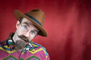 Bıyıklı, gözlüklü, kahverengi şapkalı ve kırmızı arkaplanlı klasik hippi.