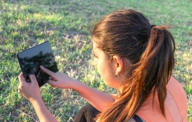Kafkasyalı genç bir kadının parkta çimlerde oturup tablete dokunuşunun görüntüsü