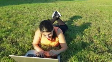 Beyaz kadının parka uzanmış, spor kıyafetleri içinde dizüstü bilgisayar kullanırken çekilmiş videosu.