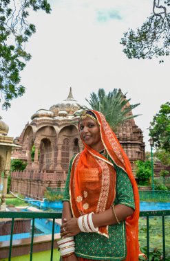 Arka planda harabelerle poz veren güzel yüzlü ve turuncu elbiseli Hintli kadın. Rajasthan, Hindistan.