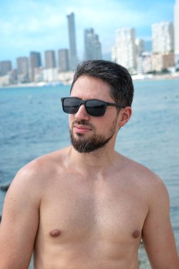 Plajda güneş gözlükleriyle gülümseyen genç İspanyol manken arka planda güzel mavi gökyüzü olan kameraya bakıyor.