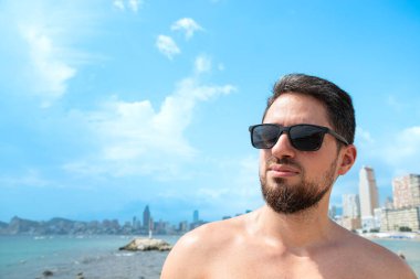 Plajda güneş gözlükleriyle gülümseyen genç İspanyol manken arka planda güzel mavi gökyüzü olan kameraya bakıyor.
