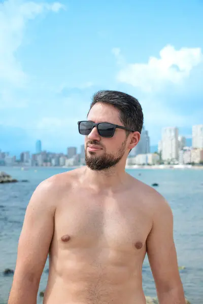 Young Hispanic man model smiling with sunglasses on beach looking to camera with beautiful blue sky in background
