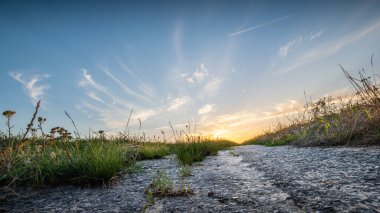 Önünde çimenler, gökte bulutlar olan bir kır yolunun üzerinde gün batımı