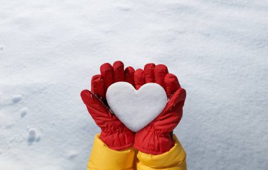 Female hand in red glove holds snow heart on snow background. Romantic winter lovers concept