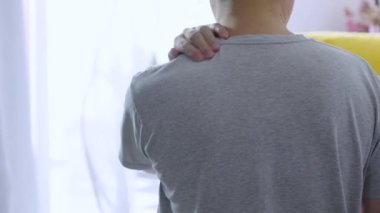 Yaşlı adam boynunu acıdan tutuyor, gri bir gömlek giyiyor.