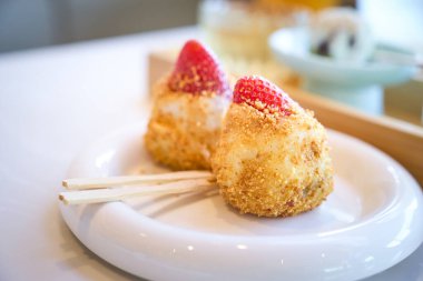 Çileklerle kaplı iki parça daifuku mochi, beyaz bir tabakta, ahşap çubuklarla tatlı bir ziyafet olarak sunulmaya hazırdır.