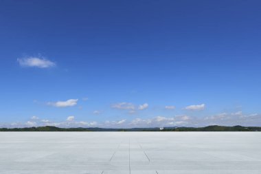 Geniş beton zemin, tüylü beyaz bulutlarla benekli, dinginlik ve açıklık hissi uyandıran huzurlu mavi bir gökyüzüne doğru uzanıyor.