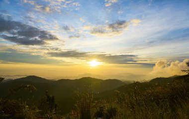 Güneş, sabahın erken saatlerinde kabarık bulutlarla bir dağ manzarasının üzerinde yükseliyor.