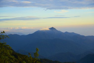 Şafak vakti bulutlarla kaplı katmanlı dağ sırası, ormanı ve gökyüzünü gösteriyor.