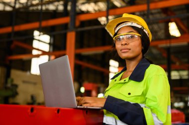 Portrait of female mechanical engineer worker in yellow hard hat and safety uniform using laptop standing at manufacturing area of industrial factory