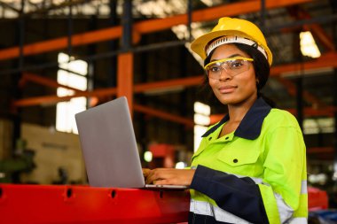 Portrait of female mechanical engineer worker in yellow hard hat and safety uniform using laptop standing at manufacturing area of industrial factory