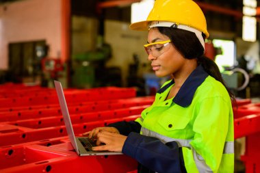 Portrait of female mechanical engineer worker in yellow hard hat and safety uniform using laptop standing at manufacturing area of industrial factory